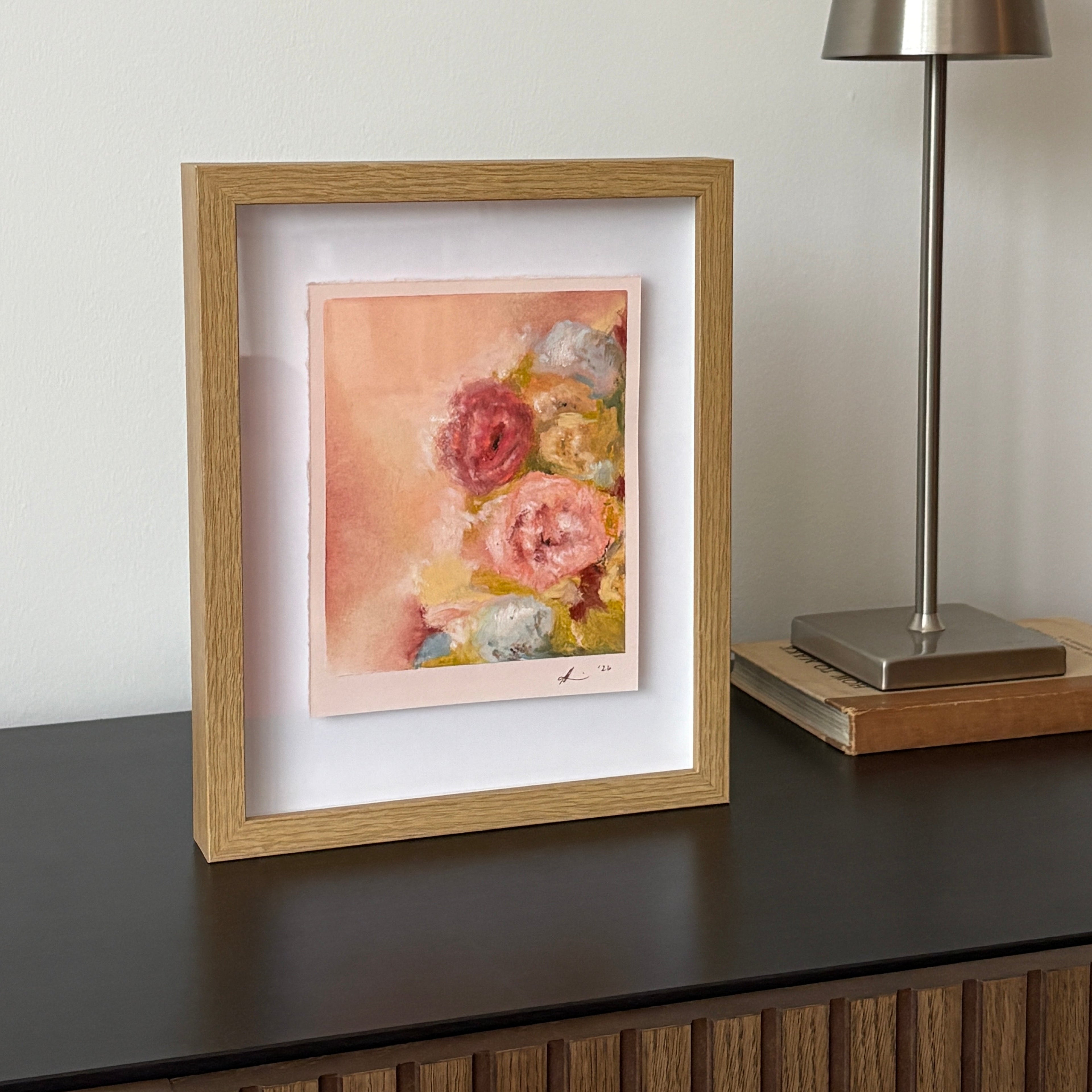 A framed floral art print with a light oak frame and white mat, displayed on a dark wooden console table next to a modern silver table lamp against a neutral off-white wall.