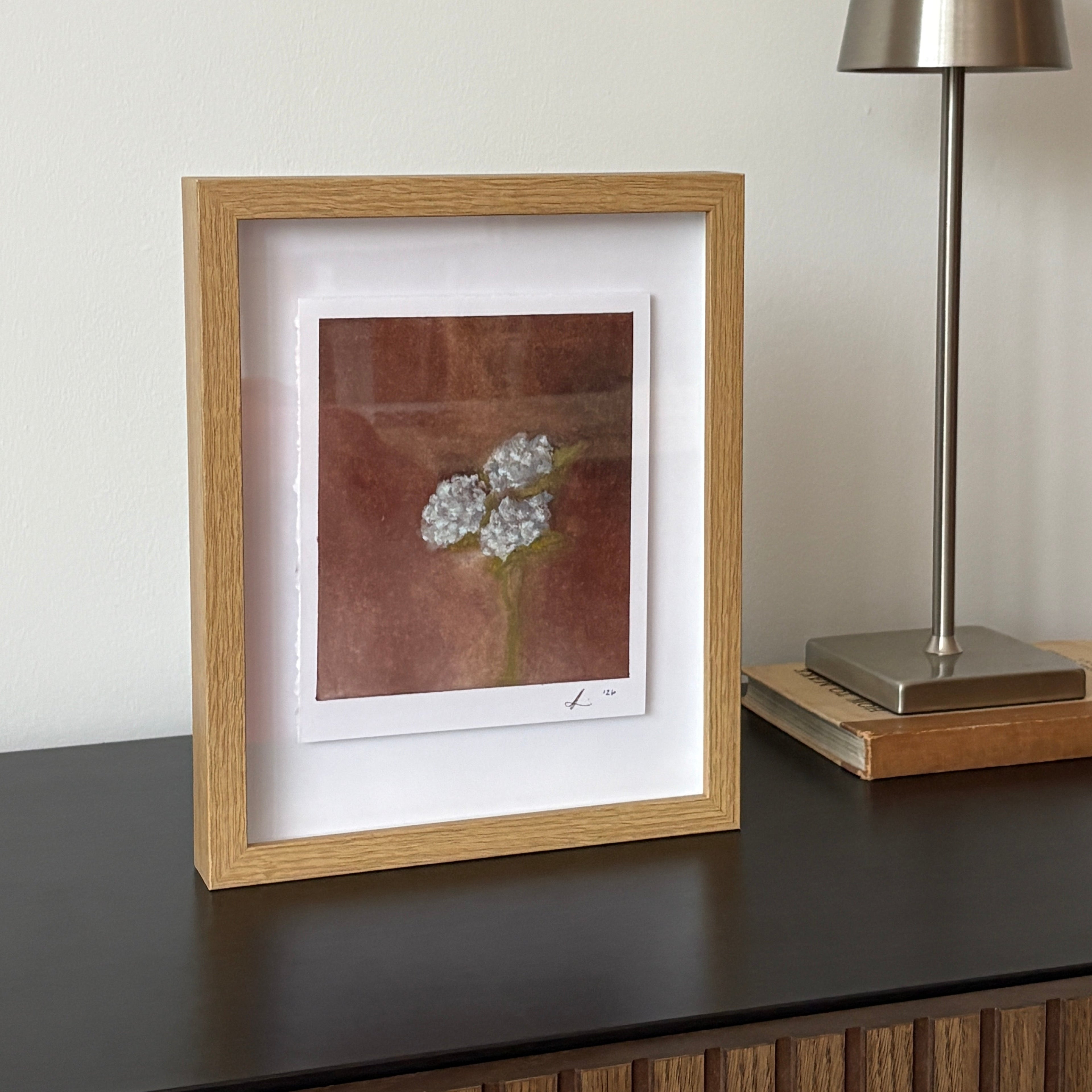 A framed botanical art print featuring white and blue flowers on a textured brown background, styled on a dark wooden console table next to a modern silver table lamp.