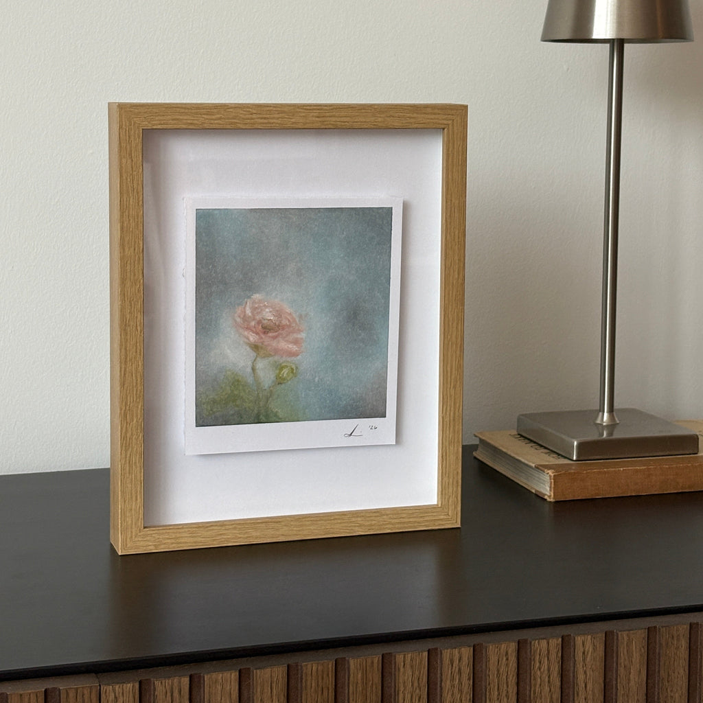 A square framed art print featuring a soft pink rose on a textured blue-gray background, set in a light oak frame with a white mat. The frame is shown next to a modern silver wall lamp.