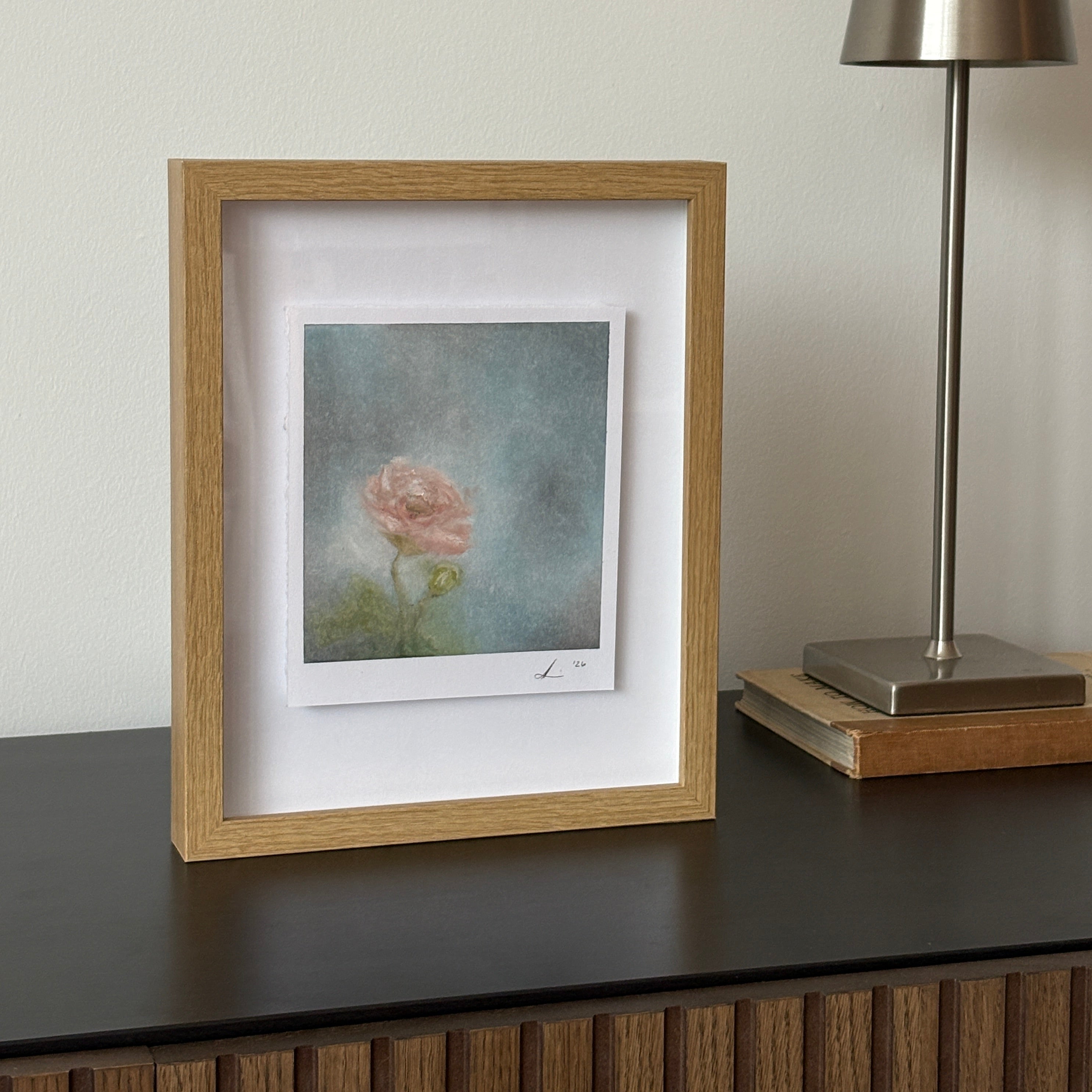 A square framed art print featuring a soft pink rose on a textured blue-gray background, set in a light oak frame with a white mat. The frame is shown next to a modern silver wall lamp.