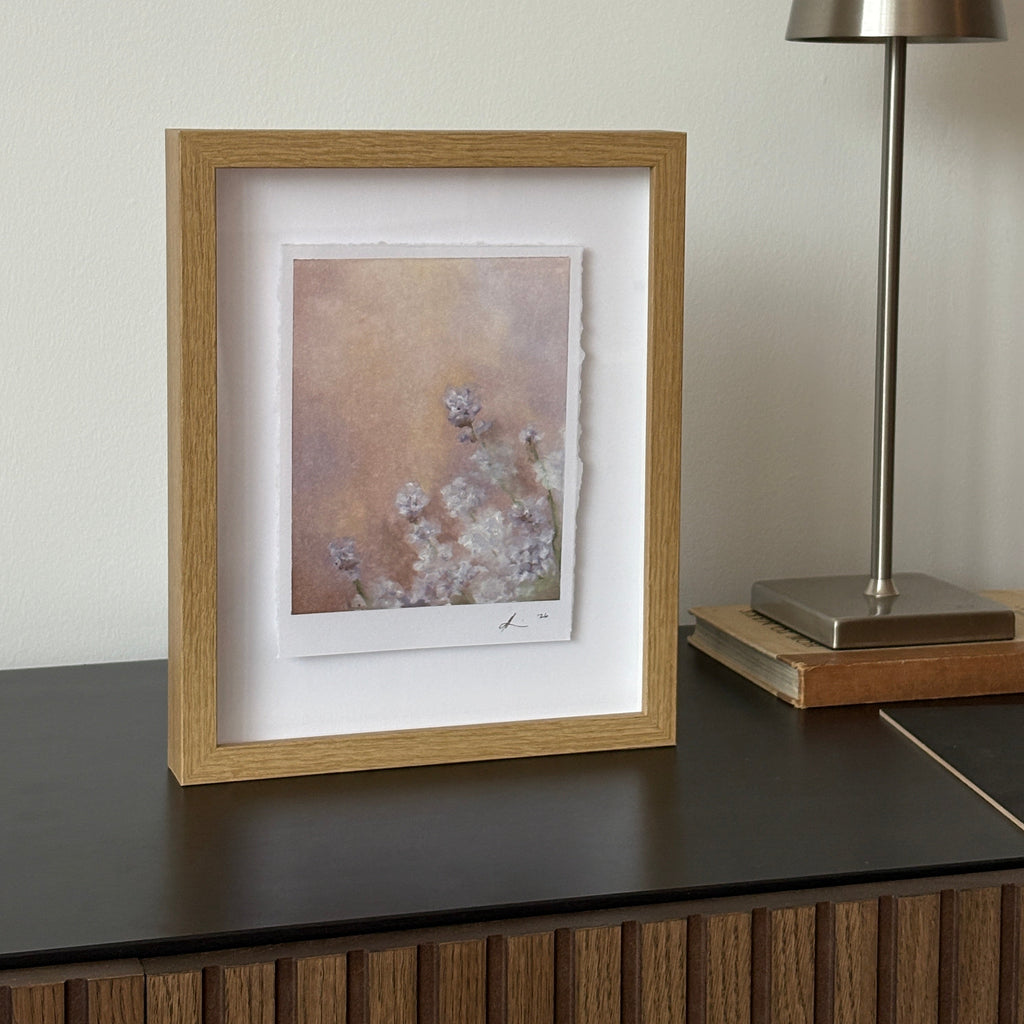 A square framed art print with a light oak wood frame and white mat, featuring a soft abstract floral design in muted pink, purple, beige, and gray tones, shown hanging on a white wall below a modern silver wall lamp.