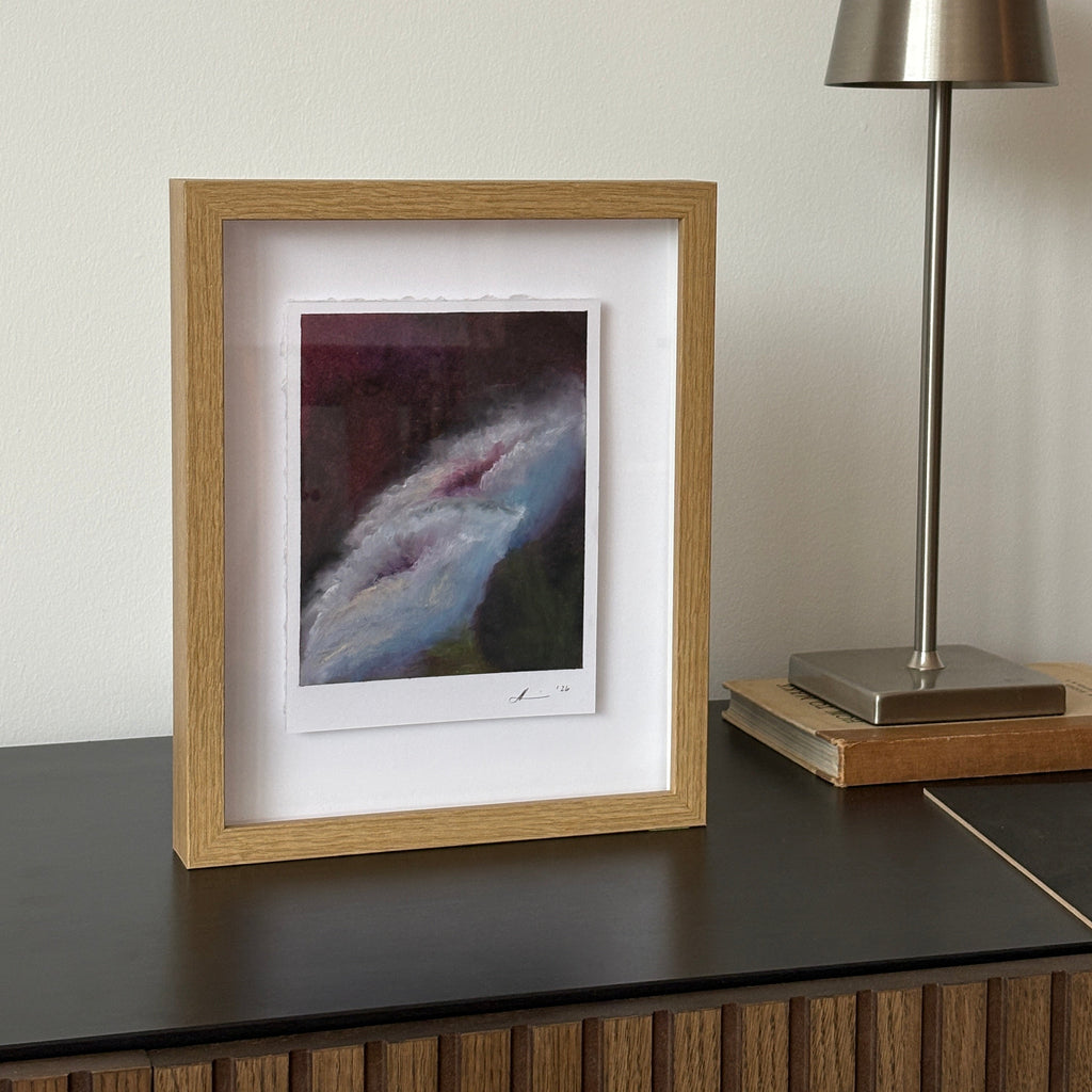 A framed abstract art print with a light oak wood frame and white mat, hanging on a white wall below a modern silver wall lamp.