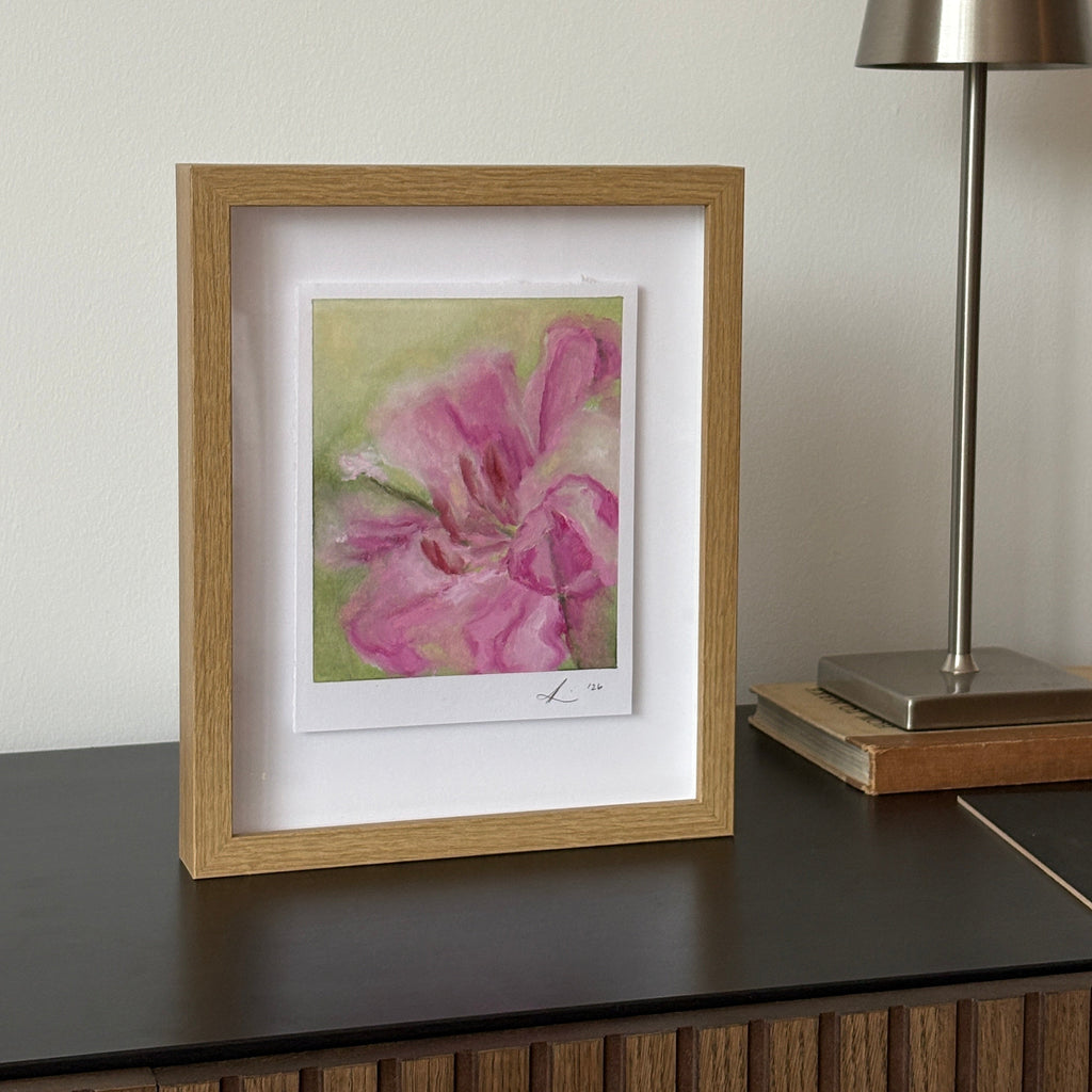 A framed watercolor painting of a pink flower in a light oak frame with a white mat, hanging on a white wall below a modern silver wall lamp.