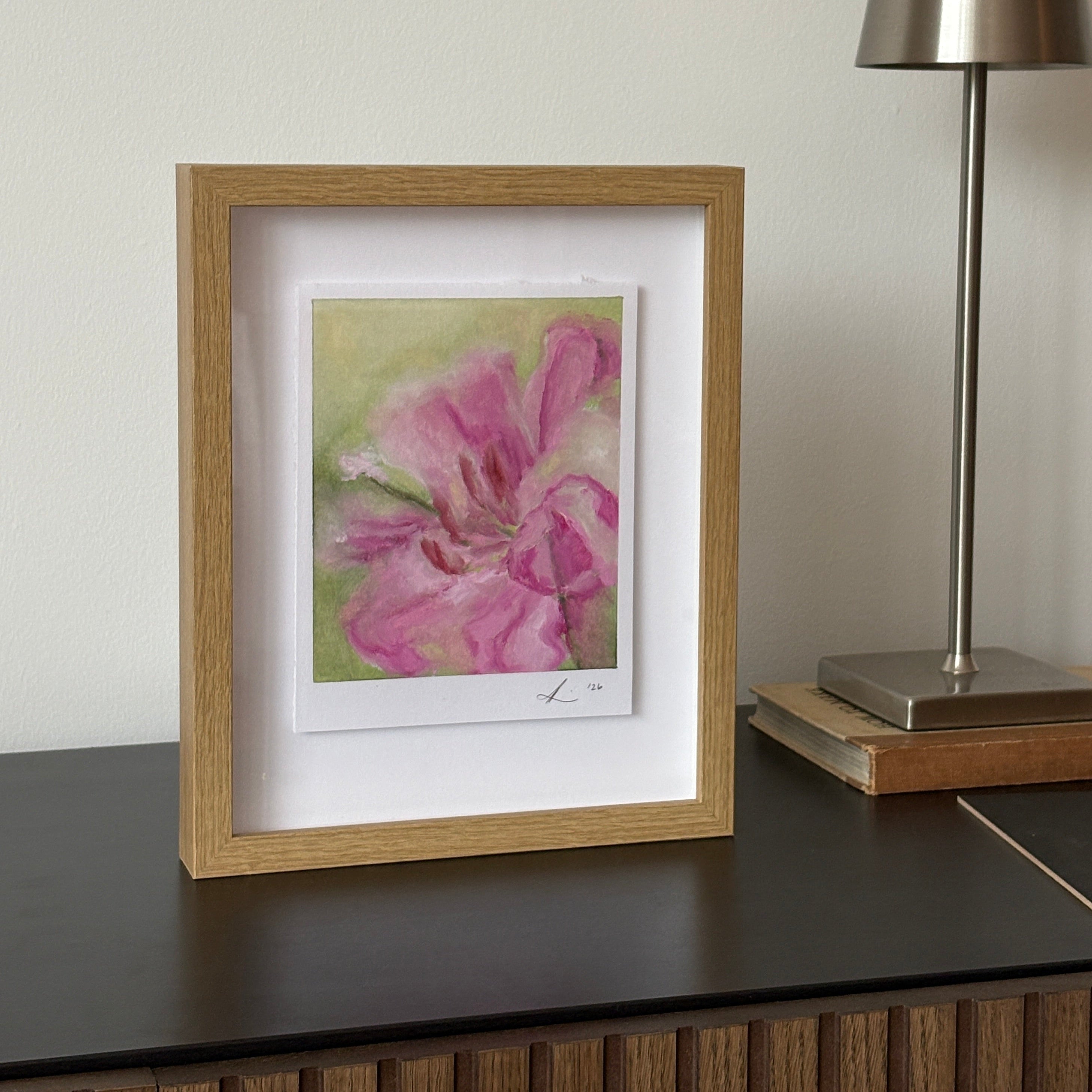 A framed watercolor painting of a pink flower in a light oak frame with a white mat, hanging on a white wall below a modern silver wall lamp.