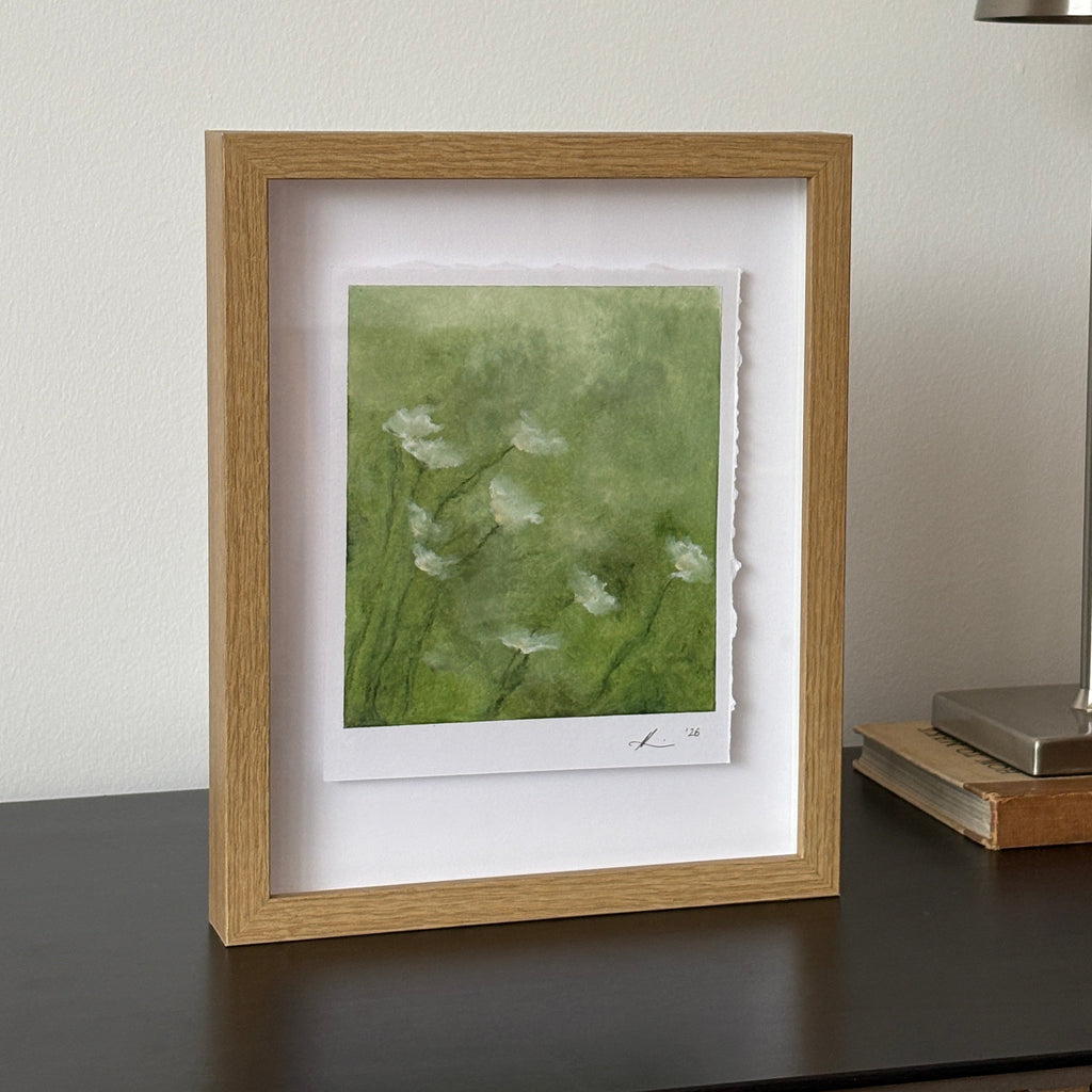 A framed art print featuring a green watercolor background with white wildflower silhouettes, housed in a light oak frame with a white mat, shown hanging on a white wall next to a modern silver wall lamp.