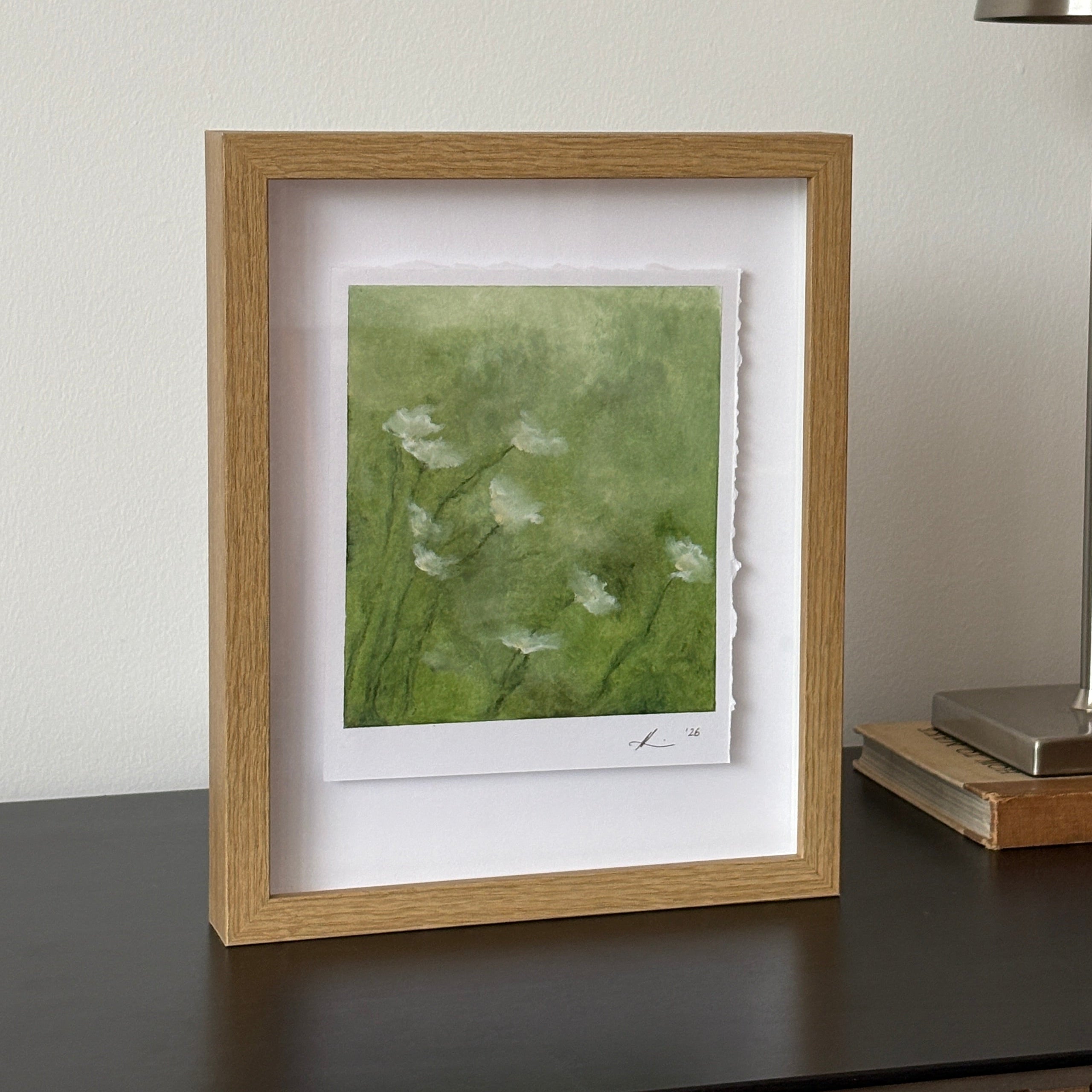 A framed art print featuring a green watercolor background with white wildflower silhouettes, housed in a light oak frame with a white mat, shown hanging on a white wall next to a modern silver wall lamp.