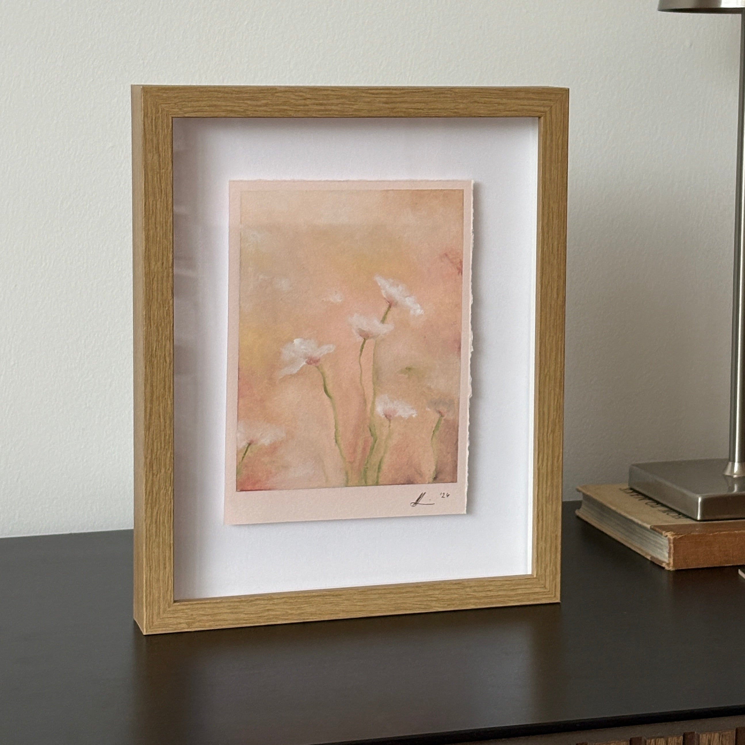A framed art print featuring a soft, abstract flowers in pastel peach, pink, and beige tones with white petals, housed in a light oak frame with a white mat. The frame is shown beside a modern silver wall lamp.