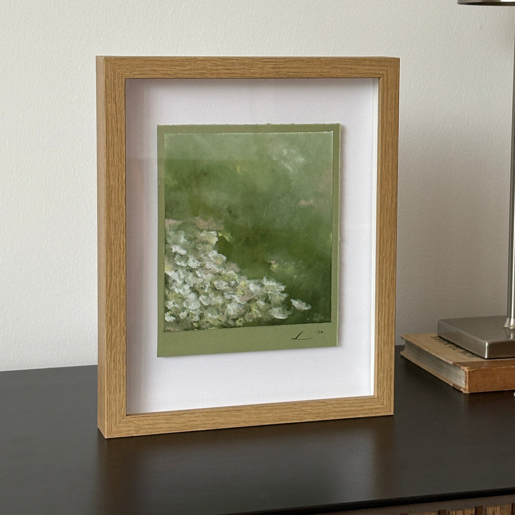 A framed art print featuring a green background with white floral details, housed in a light oak frame with a white mat, shown hanging on a white wall below a modern silver wall lamp.
