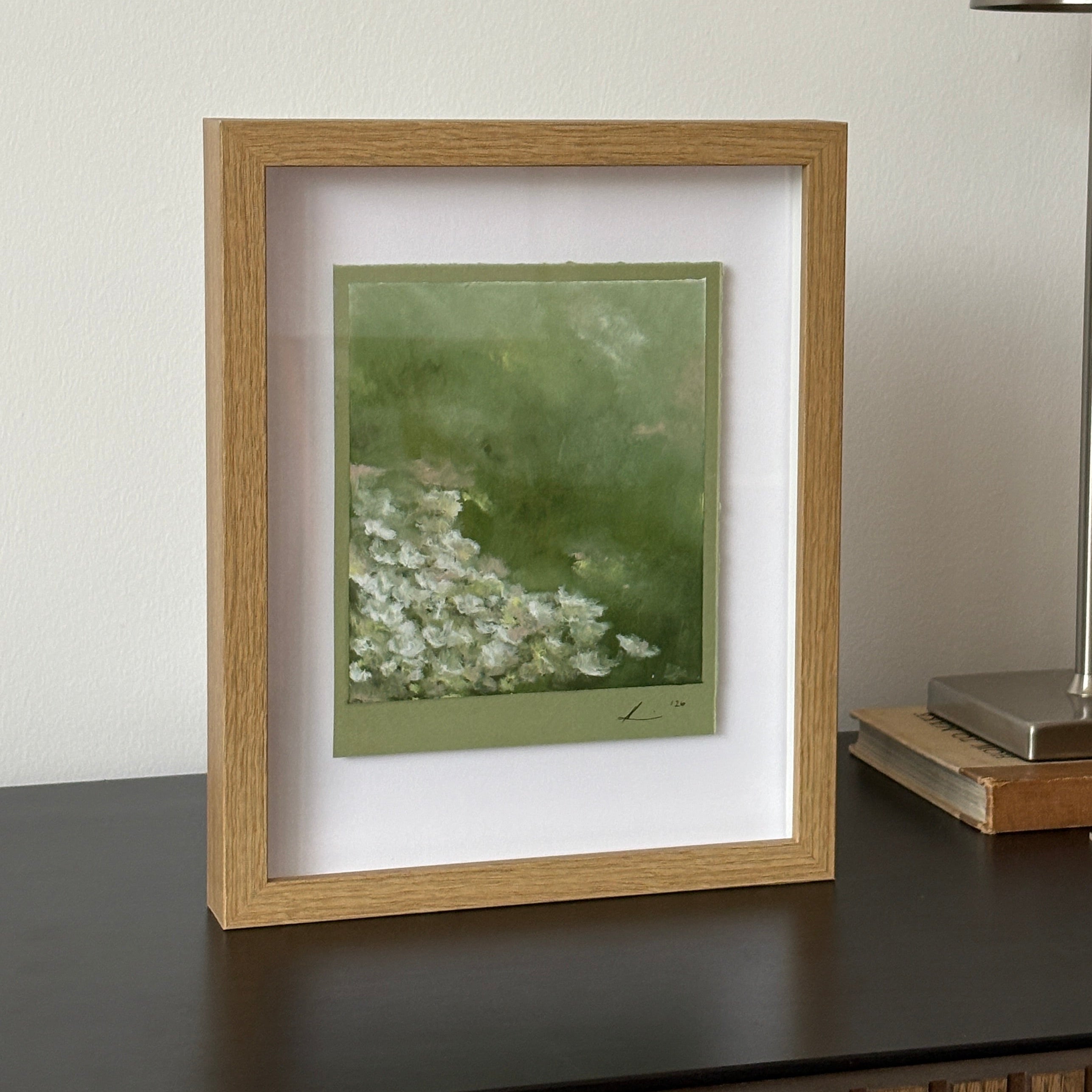 A framed art print featuring a green background with white floral details, housed in a light oak frame with a white mat, shown hanging on a white wall below a modern silver wall lamp.