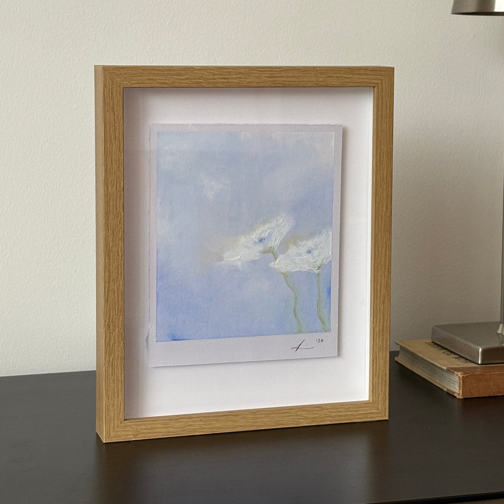 A small framed art print featuring a blue sky with white clouds, housed in a light oak frame with a white mat, shown hanging on a white wall below a modern silver wall lamp.