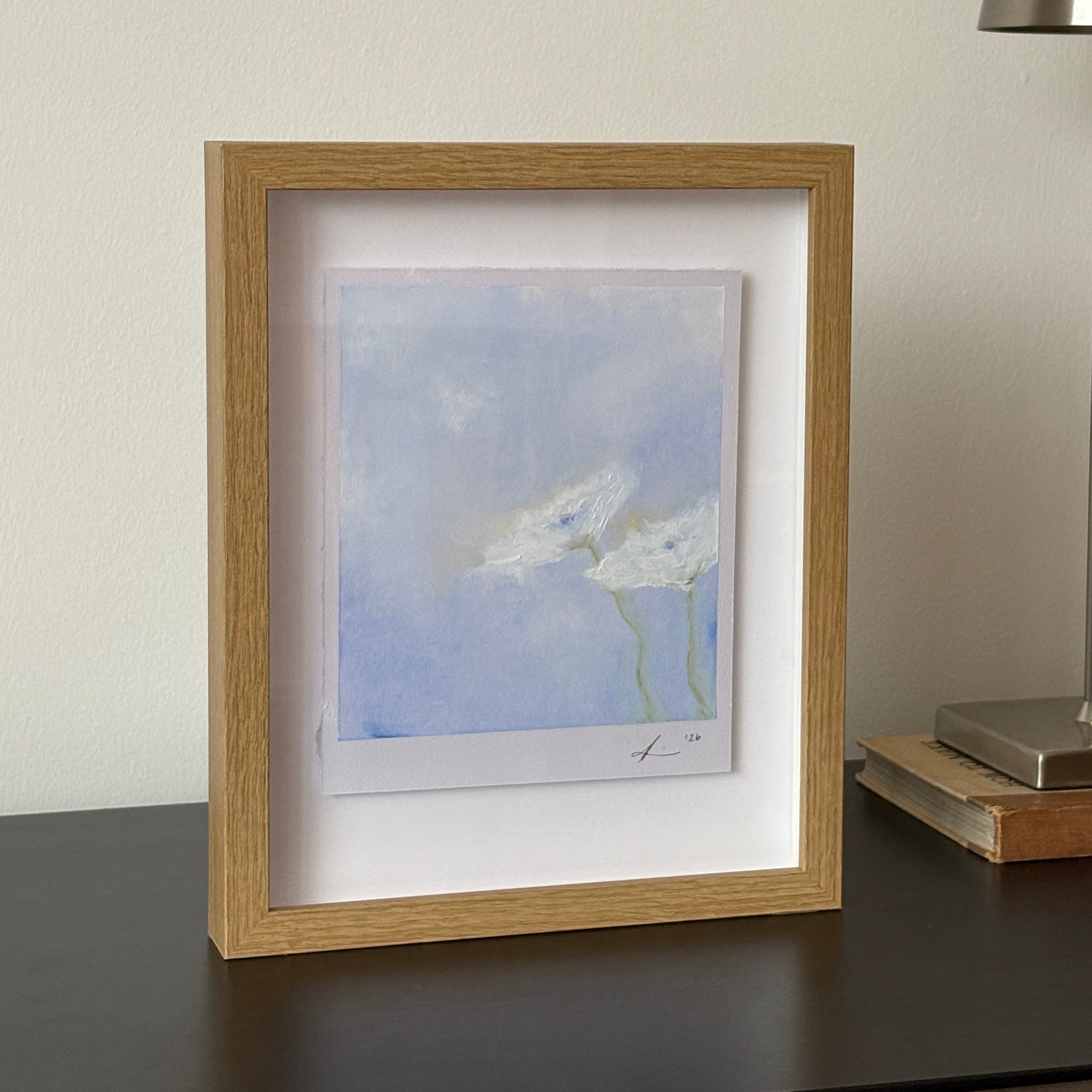 A small framed art print featuring a blue sky with white clouds, housed in a light oak frame with a white mat, shown hanging on a white wall below a modern silver wall lamp.