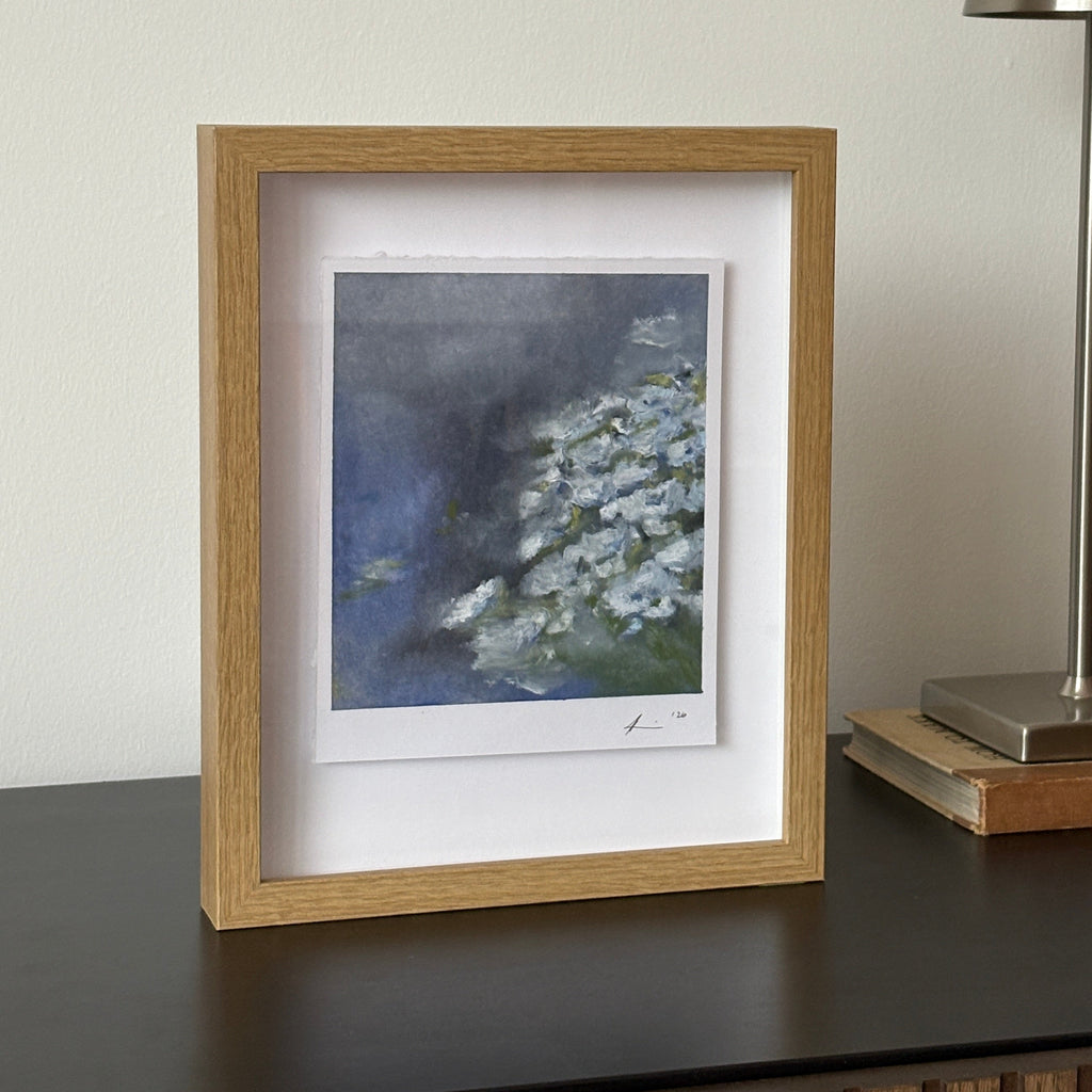 A framed art print featuring a blue and white floral painting, styled on a dark gray console table next to a modern silver wall lamp.