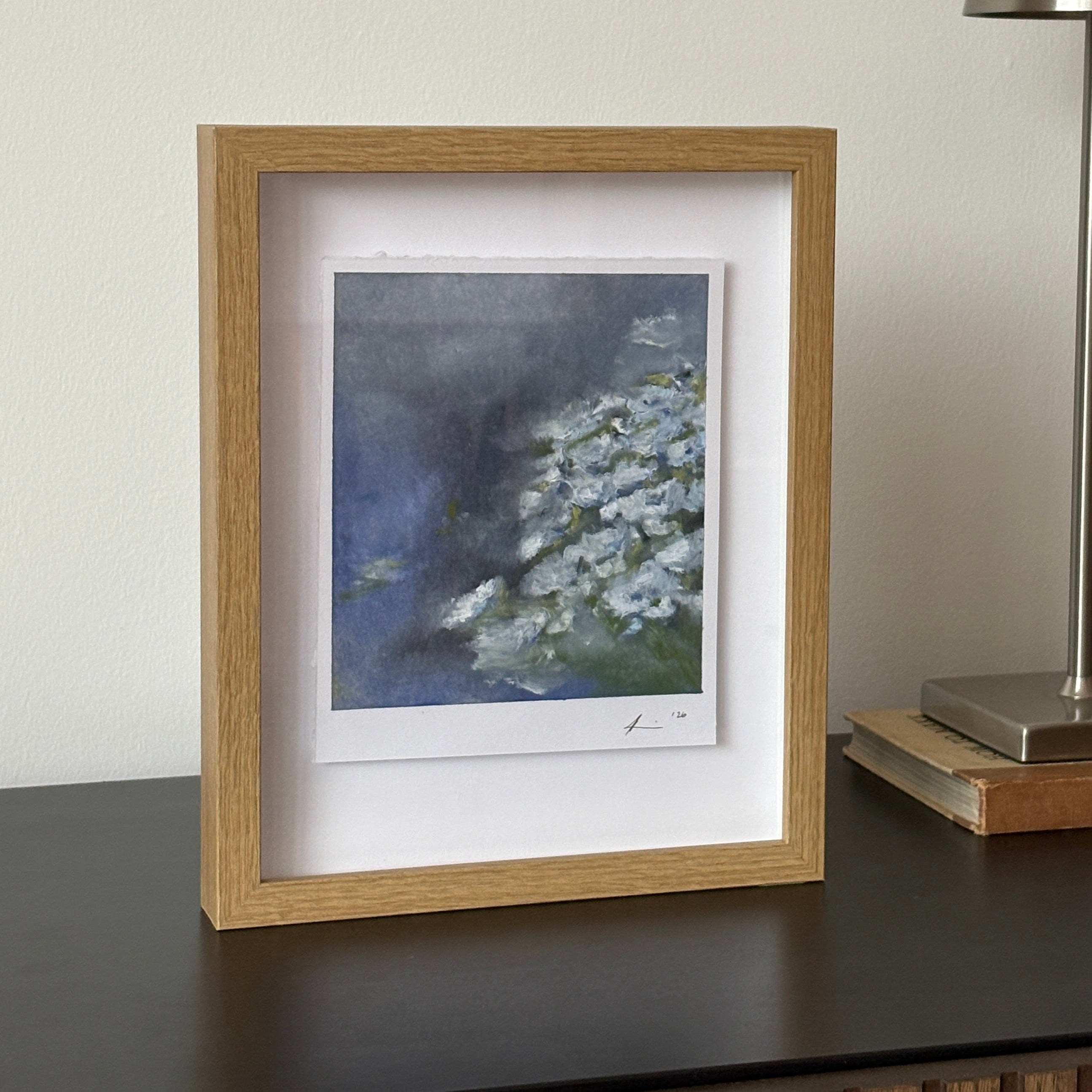 A framed art print featuring a blue and white floral painting, styled on a dark gray console table next to a modern silver wall lamp.