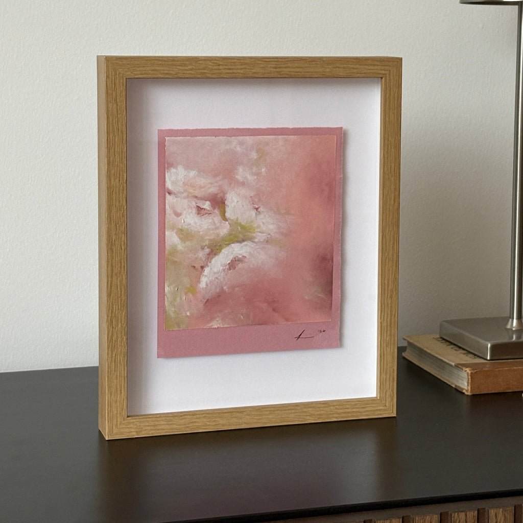 A framed abstract art print featuring pink and white textured marks, housed in a light oak frame with a white mat, shown hanging on a shelf a modern silver wall lamp.