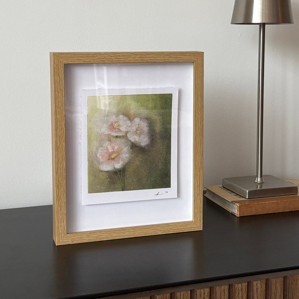 A framed floral art print with a light oak frame and white mat, displayed on a dark wooden sideboard next to a modern silver table lamp.